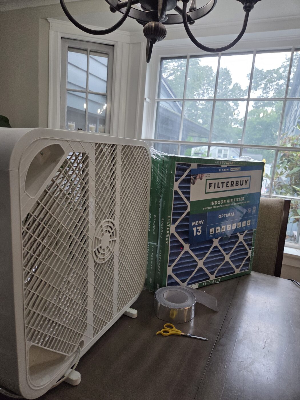 A box fan, a pack of MERV 13 filters, foil tape and scissors laid out by a window.